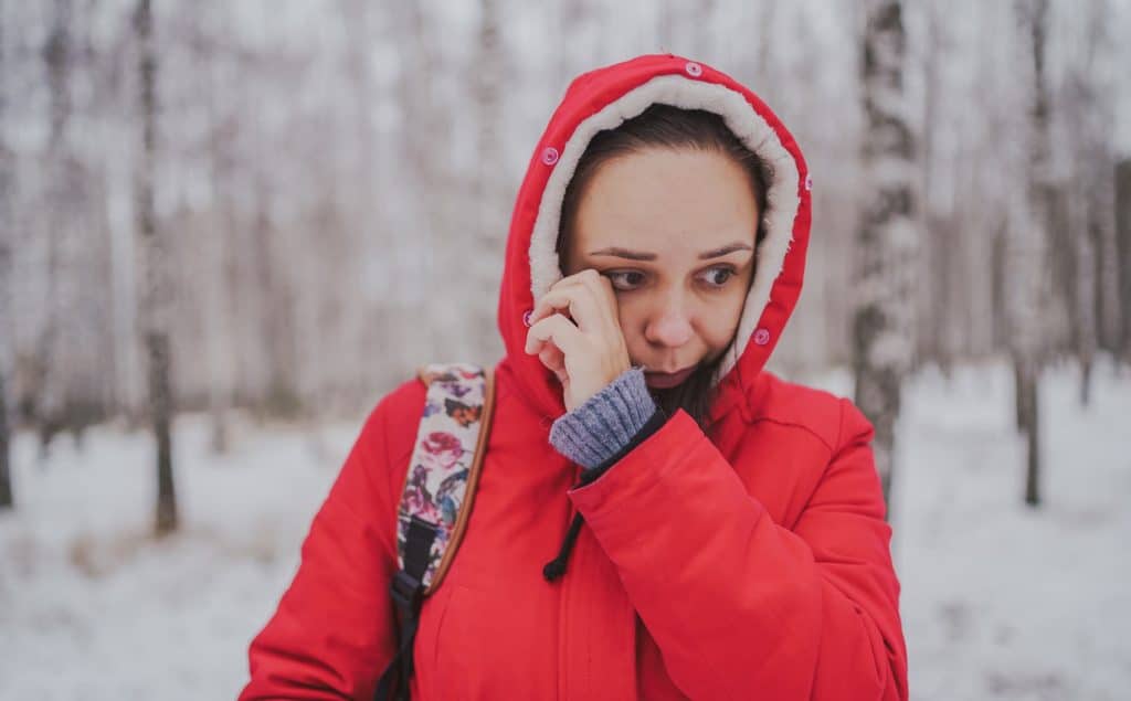 A young woman in a red coat rubs her eyes in a snowy forest.